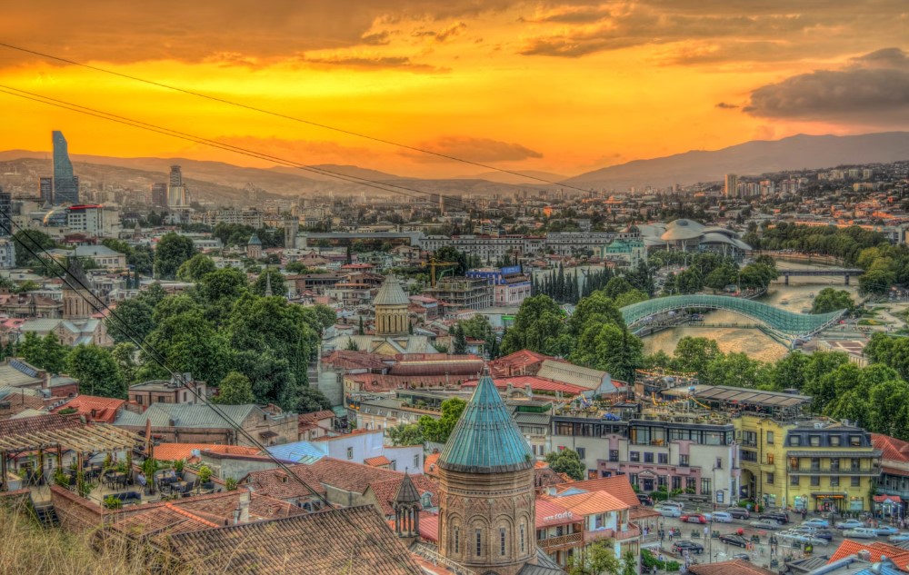 Sunset glow over Tbilisi’s historic rooftops, the Kura River, and the modern “Bridge of Peace.”