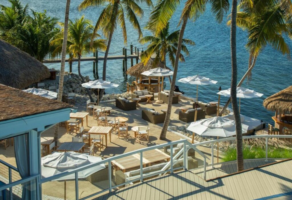 Wicker lounges shaded by palms along Bungalows Key Largo’s sunlit bayfront